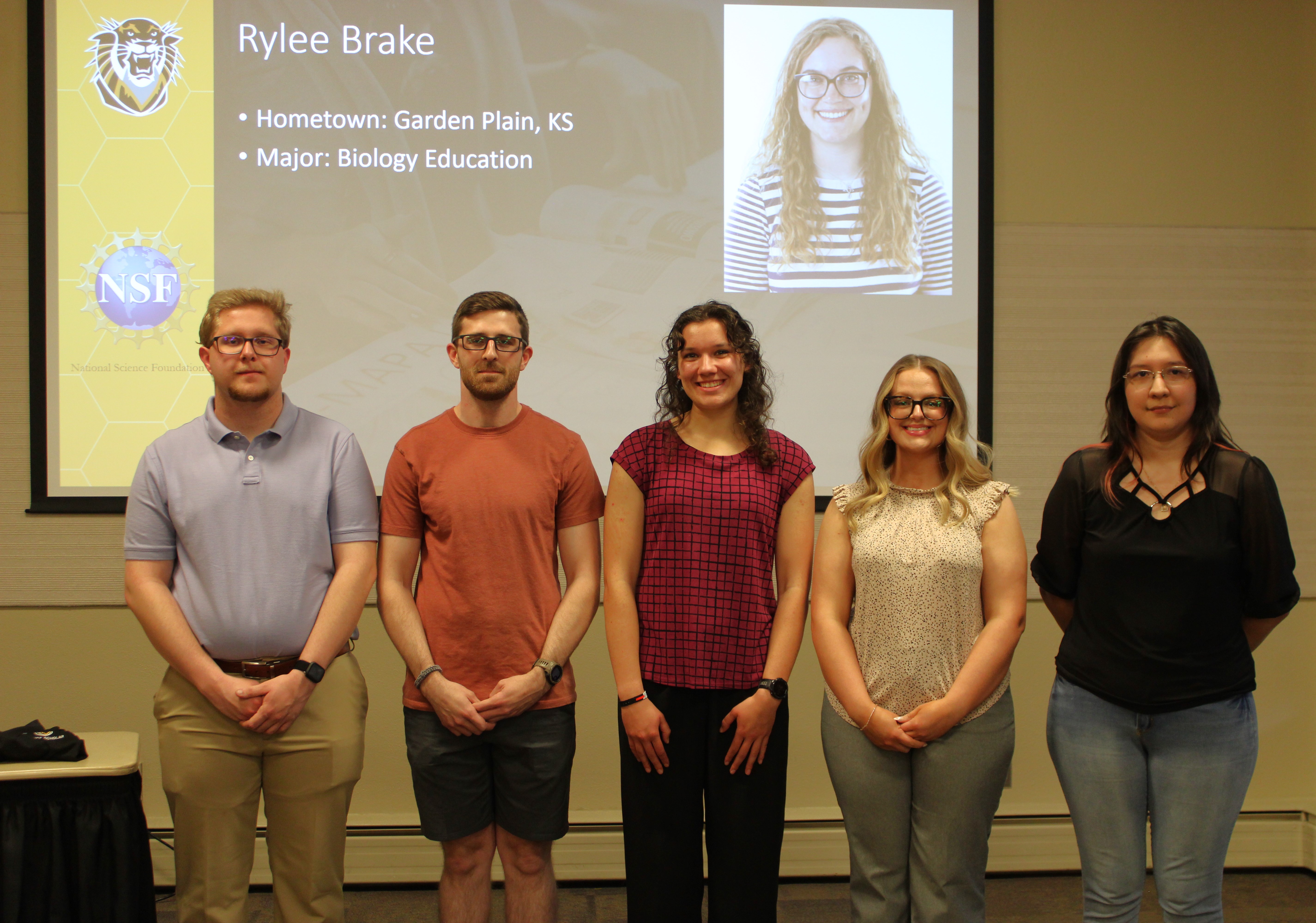2025 Noyce Scholars, left to right: Dustin Rajewski, Ryan Lawson, Ellie Van Zelfden, Jaylei Sloan, and Jazmin Contreras. Rylee Brake is shown on the projection screen.