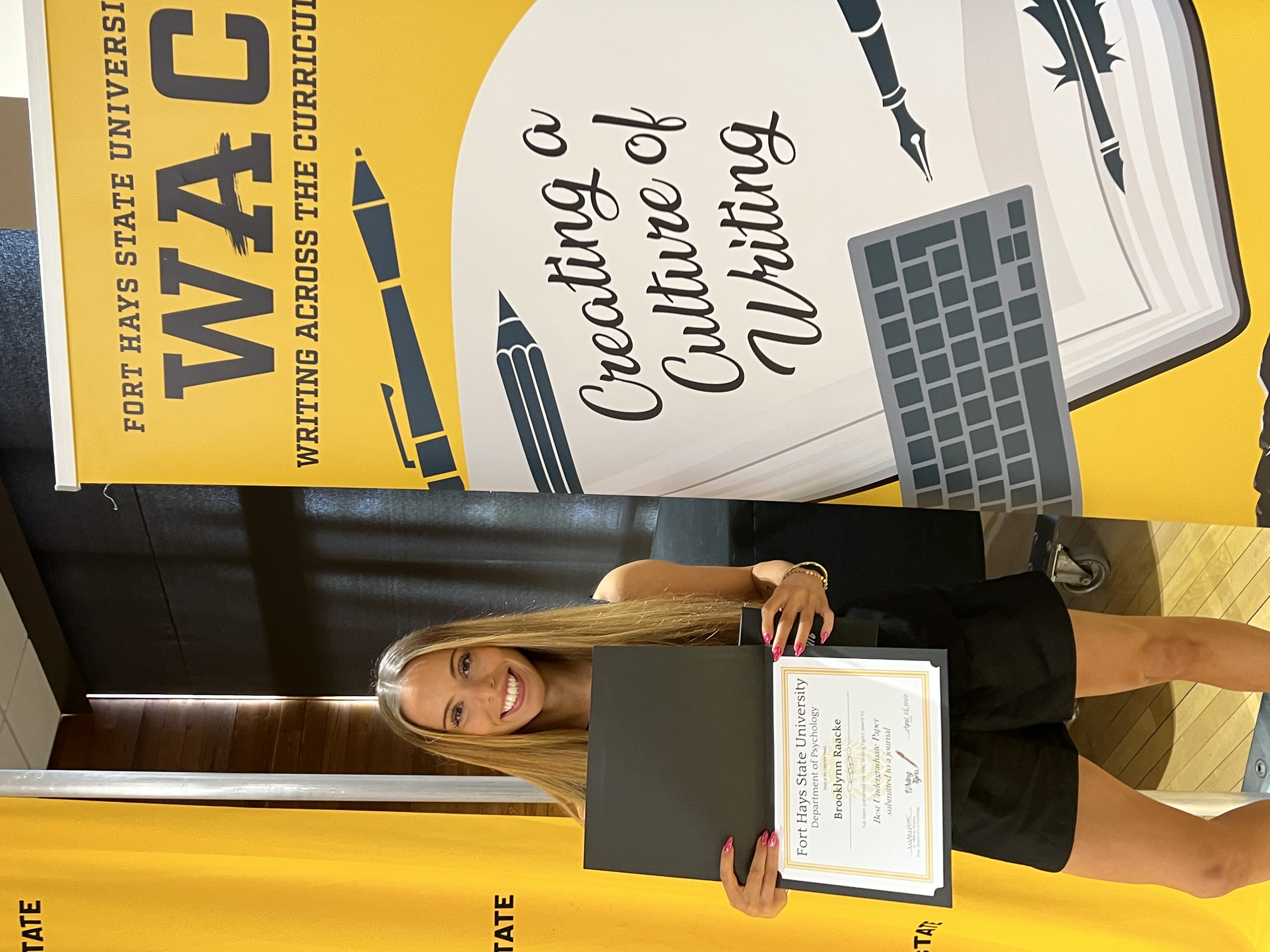 Student wearing a black dress holding award in front of WAC banner
